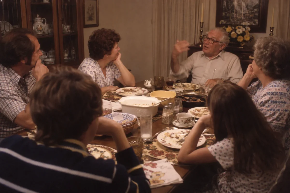 Someone telling a story at a dinner table while everyone listens, around 1980